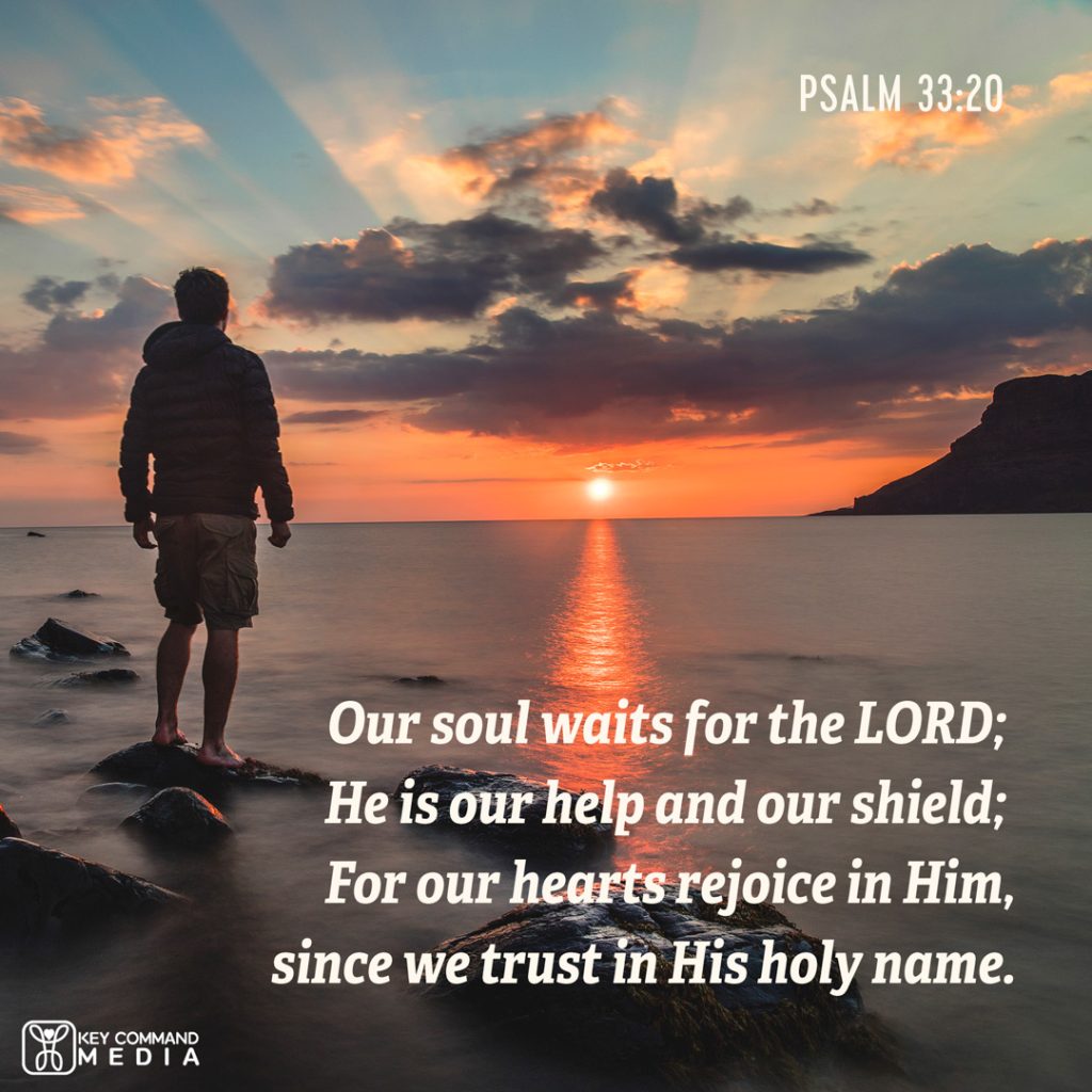 A young man in a hoodie stands at the seashore watching a sunset over the water with Psalm 33:20, "Our soul waits for the Lord; He is our help and shield; For our hearts rejoice in Him, since we trust in His holy name."
