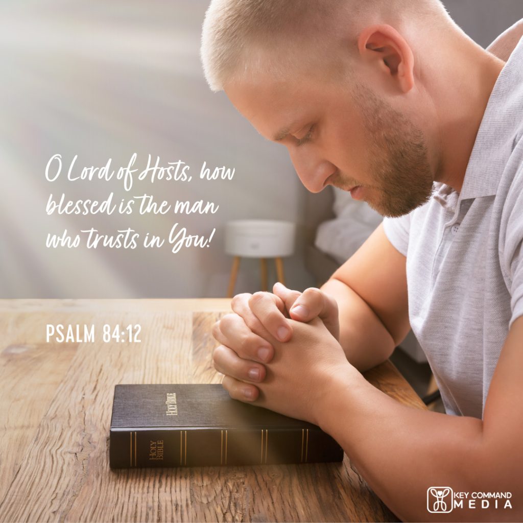 A young man prays seated at a table with his hands folded on top of a Bible with Psalm 84:12, "O Lord of Hosts, how blessed is the man who trusts in You!"