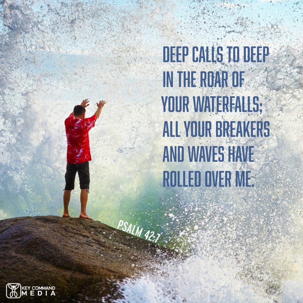 A man is getting drenched by crashing waves as he stands near the edge of a large rocky shore with Psalm 42:7, "Deep calls to deep in the roar of your waterfalls; all your breakers and waves have rolled over me."