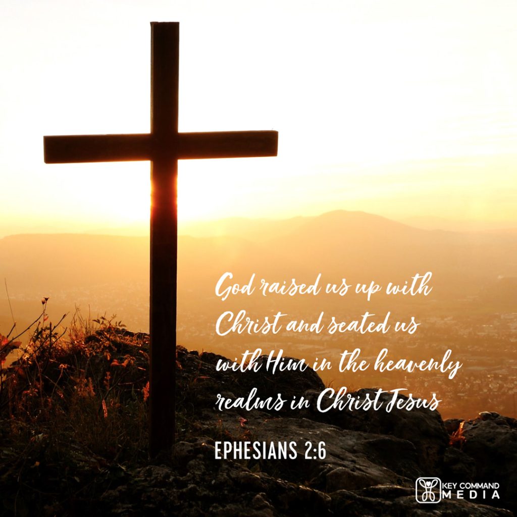 A cross is atop a high hill as the yellow sun rises with Ephesians 2:6, "God raised us up with Christ and seated us with Him in the heavenly realms in Christ Jesus."