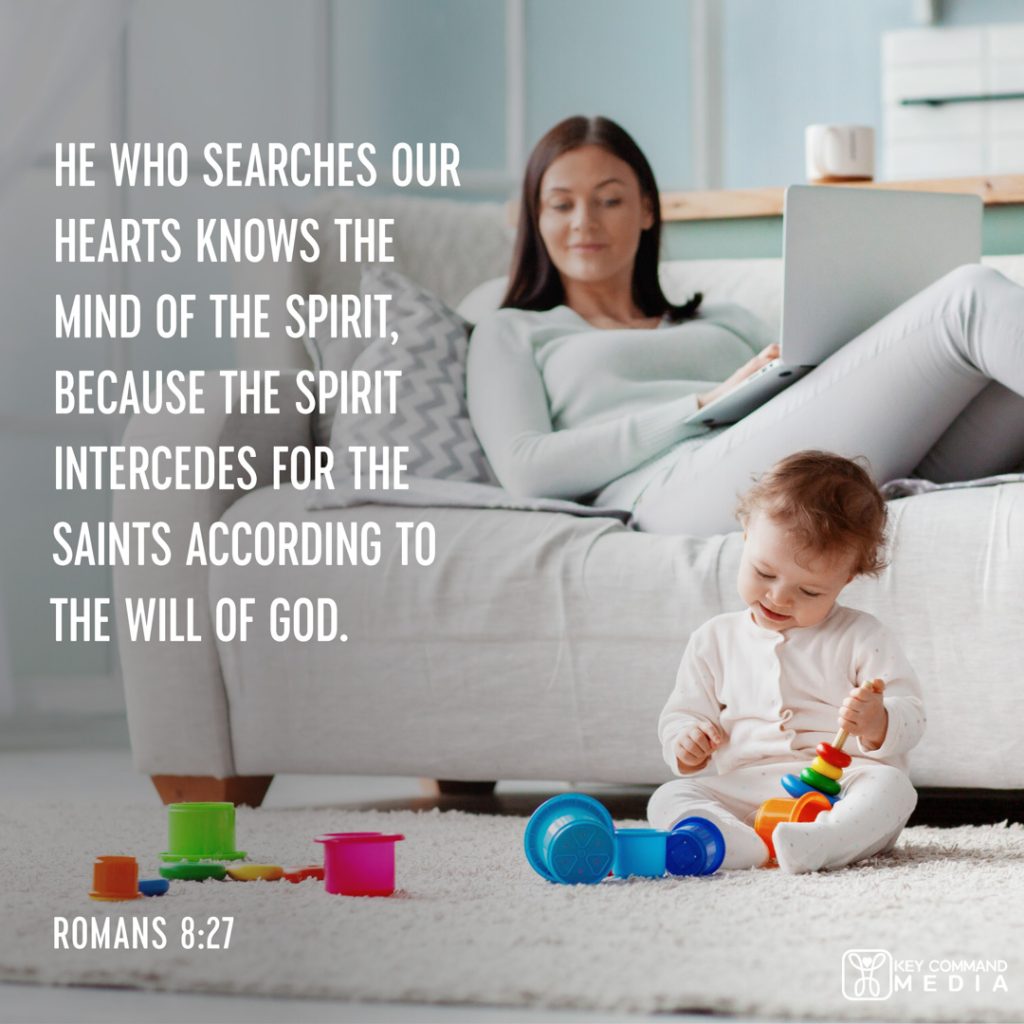 A mother using a laptop on a couch watches over a toddler playing with toys right below her with Romans 8:27, "He who searches our hearts knows the mind of the Spirit, because the Spirit intercedes for the saints according to the will of God."