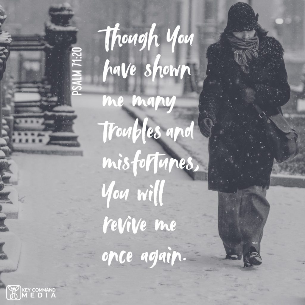 A woman bundled up in winter clothes walks down a snowy city sidewalk with snow falling with Psalm 71:20, "Though You have shown me many trouble and misfortunes, You will revive me once again."