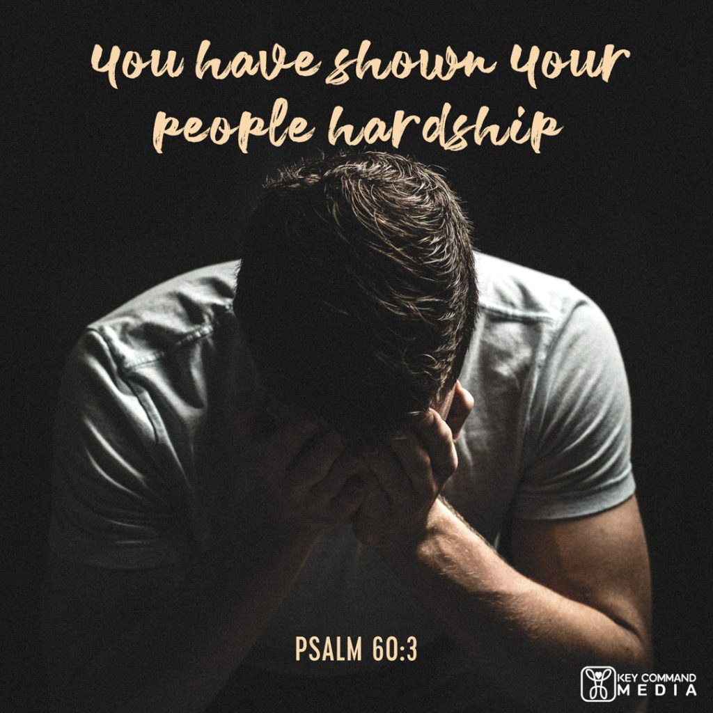 A young man in shadow and black background has his head down with his hands over his face with Psalm 60:3, "You have shown Your people hardship."