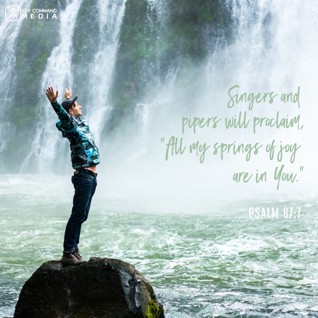 A smiling man stands on a large boulder with arms held high in front of a waterfall and springs with Psalm 87:7, "Singers and piper will proclaim, "All my springs of joy are in You."
